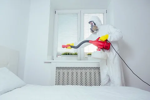 Ein Schädlingsbekämpfer in Schutzanzug, Handschuhen und Atemmaske führt eine Bettwanzenbekämpfung im Schlafzimmer durch. Er richtet ein rotes Gerät auf das Bett. Der Raum ist hell und modern eingerichtet, mit einem Fenster im Hintergrund.